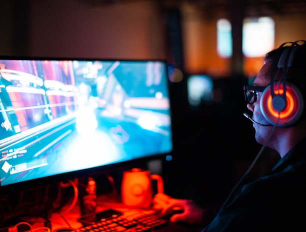 A person wearing headphones is sitting in front of a computer monitor. The screen displays a vibrant, illuminated video game scene, while the person appears focused on gaming. The ambient lighting is dark with red and blue tones, and a backlight from the monitor highlights the headphones.