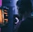 A person wearing headphones is engaged in gaming on a computer, with a large screen displaying a video game scene, showing a wooden building and a path.