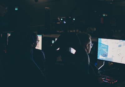 Several individuals are sitting in front of computer screens in a dimly lit room, likely engaging in gaming or working on tasks. The glow of the monitors illuminates their faces, creating a focused and immersive atmosphere. The surroundings suggest a setting like a gaming event or a LAN party.