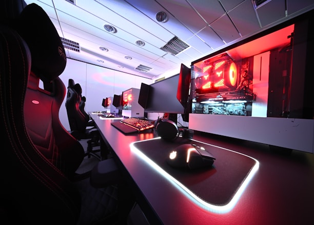 A row of high-end gaming computers on sleek desks, featuring RGB lighting and gaming accessories. The computers are housed in transparent cases with visible components, emitting a red glow. Comfortable gaming chairs with ergonomic design are positioned next to the desks, and the room's ceiling is modern with recessed lighting.