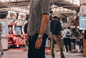 A person stands in a lively arcade setting at night, surrounded by string lights and various arcade game machines. The scene is bustling with activity as other people are visible in the background, engaging with the games or walking by. The signage reads 'Game Zone', indicating the entertainment venue.
