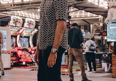 A person stands in a lively arcade setting at night, surrounded by string lights and various arcade game machines. The scene is bustling with activity as other people are visible in the background, engaging with the games or walking by. The signage reads 'Game Zone', indicating the entertainment venue.