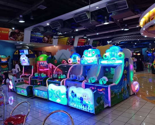 A brightly lit arcade filled with colorful gaming machines sits under ceiling lights. Neon lights illuminate various game stations, creating a playful and vibrant environment. A few red chairs are scattered around the reflective, tiled floor, and a person is observing one of the machines. The atmosphere is lively and cheerful, featuring games with cartoonish designs and engaging themes.